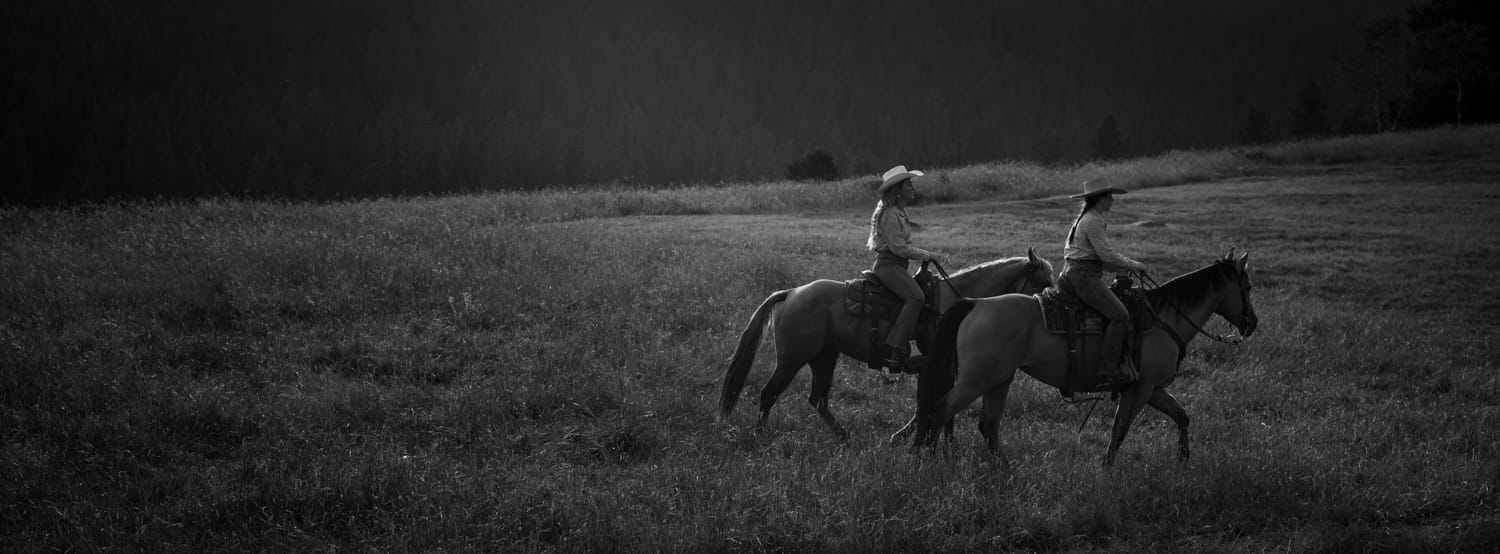 Black and white phot of two people on horseback riding through a field.