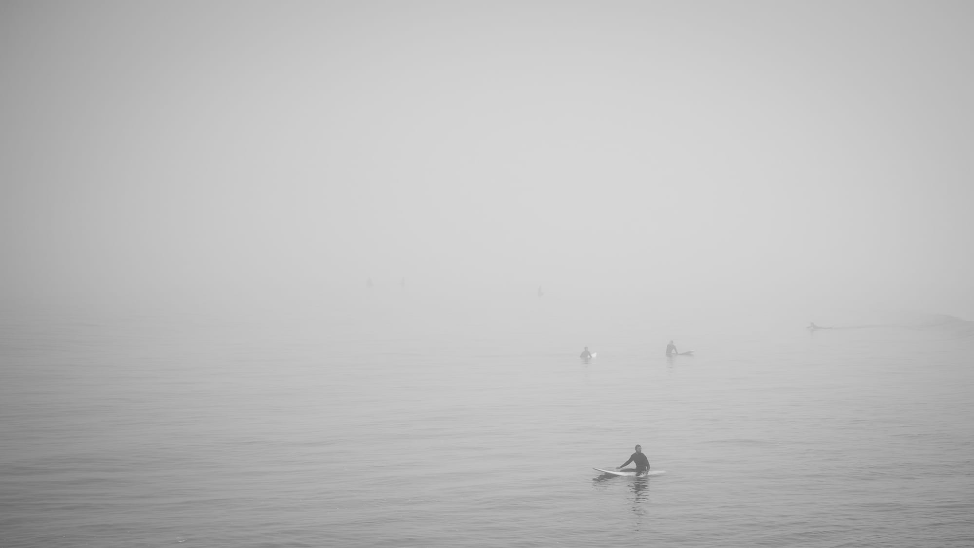 Black and white photo of three surfers sitting on their boards surrounded by a thick fog.