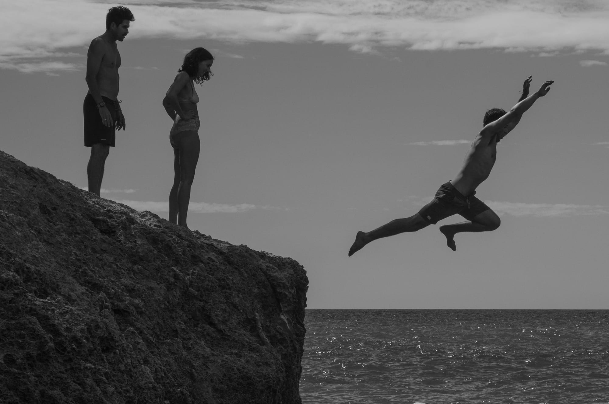 A man jumps off a cliff into the ocean while two bathers await their turn.