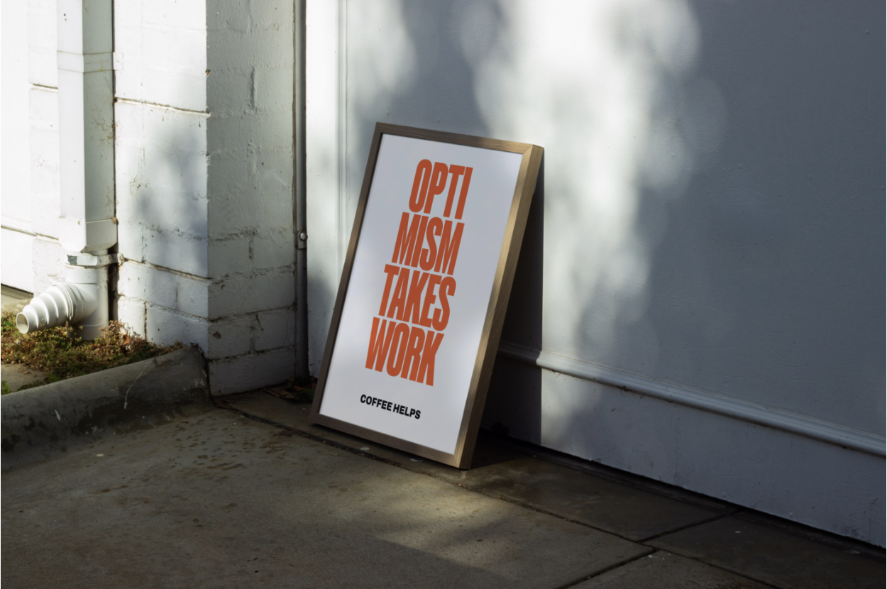 Photo of a framed poster that reads OPTIMISM TAKES WORK. COFFEE HELPS.